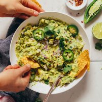 Overhead image of guacamole in a white bowl and a hand scooping some guacamole with a chip