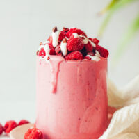 Overflowing glass of our raspberry banana smoothie topped with raspberries, coconut yogurt, and cacao nibs