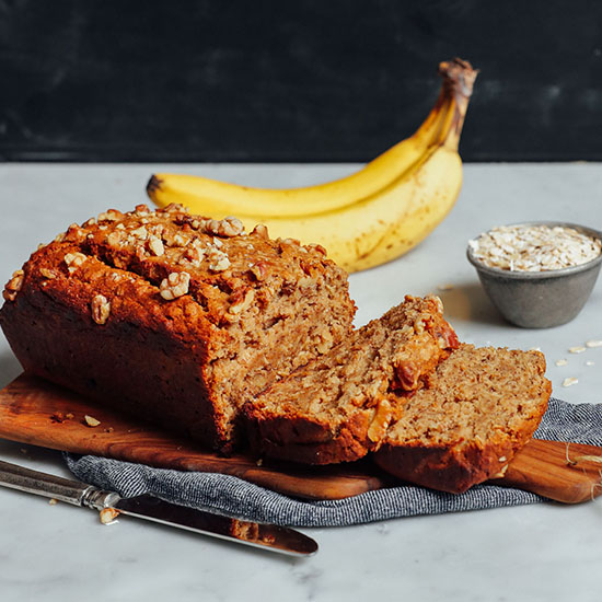 Partially sliced loaf of Vegan Gluten-Free Banana Bread with bananas and oats in the background
